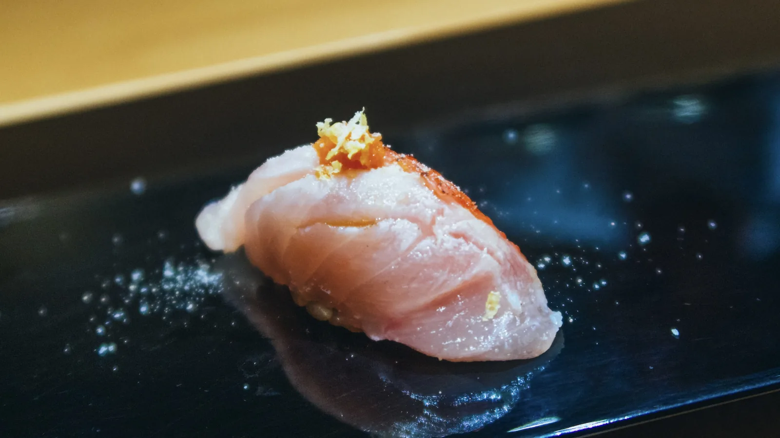 A piece of kinmedai nigiri, its skin seared and topped with yuzu zest, on a dark lacquered plate